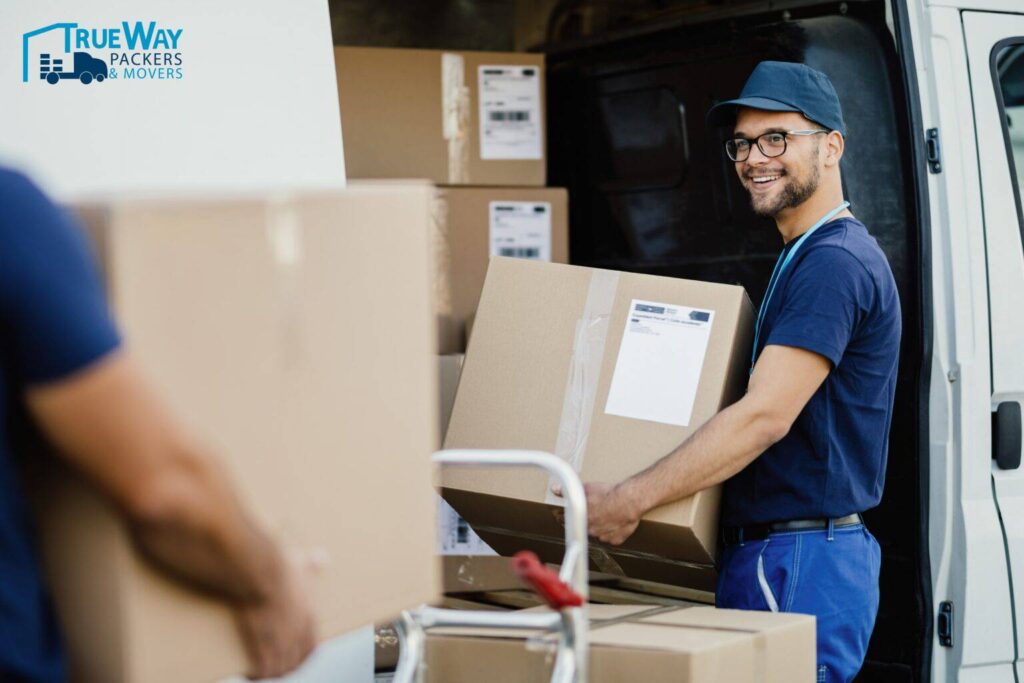 Young happy manual worker carrying cardboard boxes delivery van while communicating with his colleagues min 1536x1024 (1)